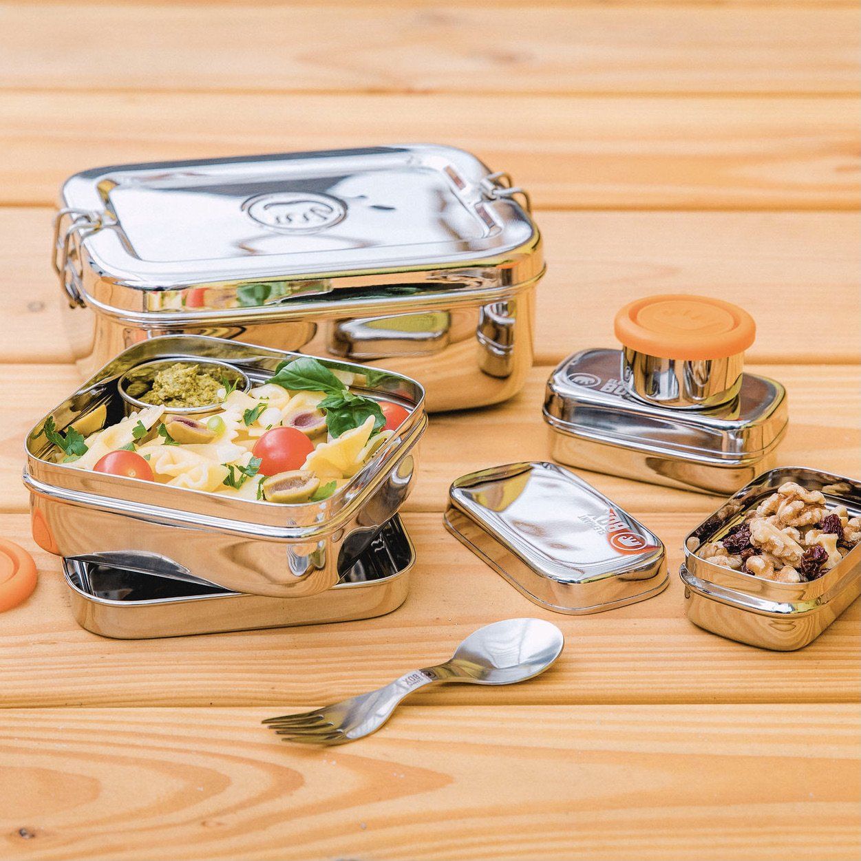 A set of stainless steel food containers including an Elephant Box 2 litre lunch box, small dressing pots with silicone lids, and a spork, all displayed on a white surface.