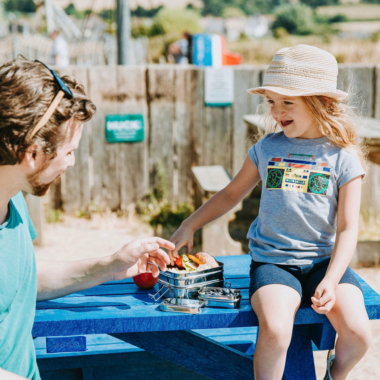a child and a man with a two tiered lunchbox and snack pod eating lunch on a picnic bench