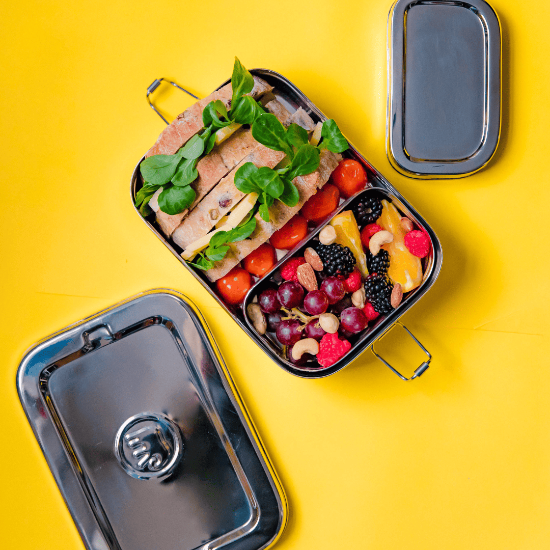 Jumbo 2 in 1 stainless steel lunch box from elephant box. box is open showing the inner snack pod containing fresh fruit and nuts. The main compartment has some chunky sandwiches & cherry tomatoes. on a bright yellow background.