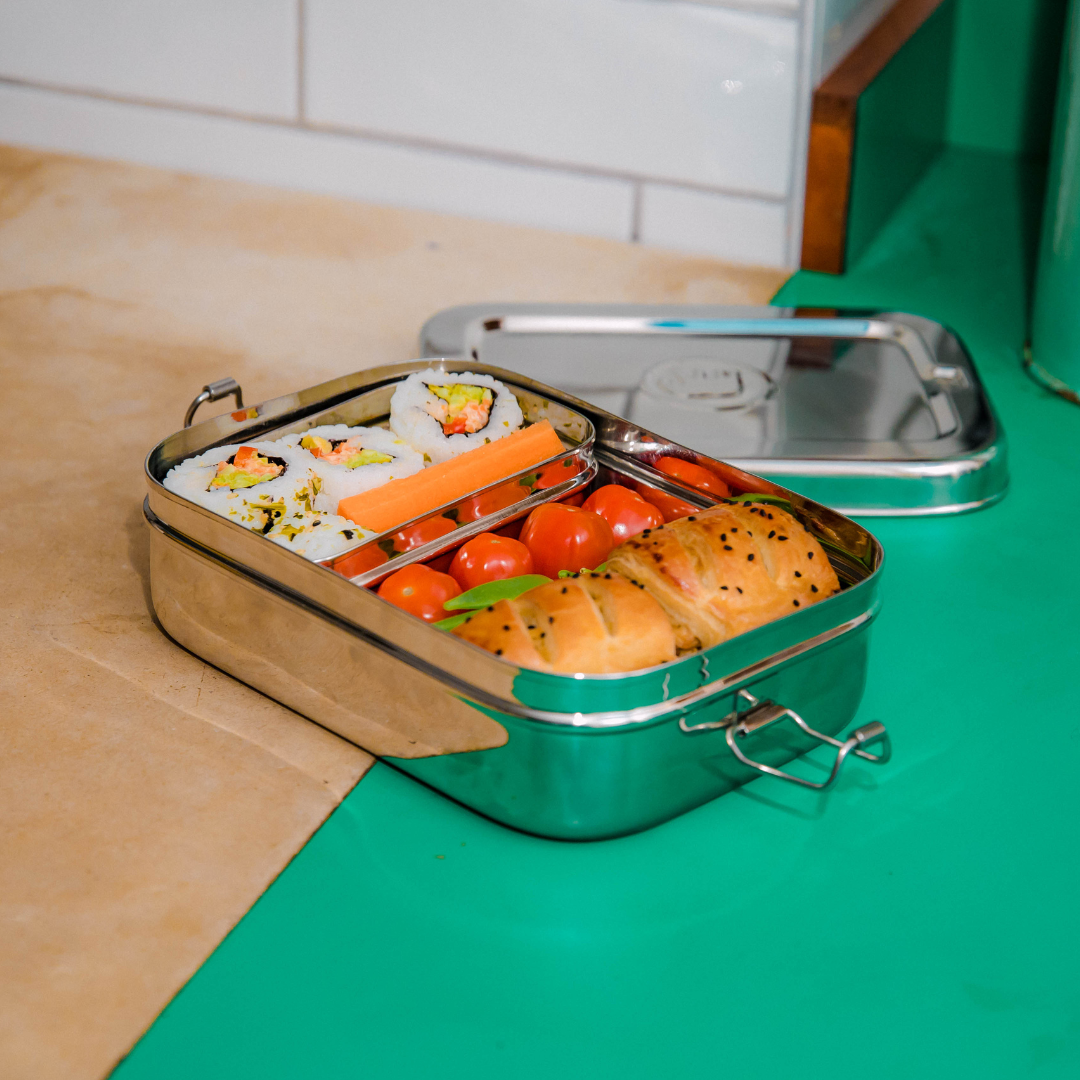 Jumbo 2 in 1 stainless steel lunch box from elephant box. box is open showing the inner snack pod containing sushi & carrot sticks.The main compartment has some cherry tomatoes, mange tout & cheese or sausage rolls. box is on a green work top.