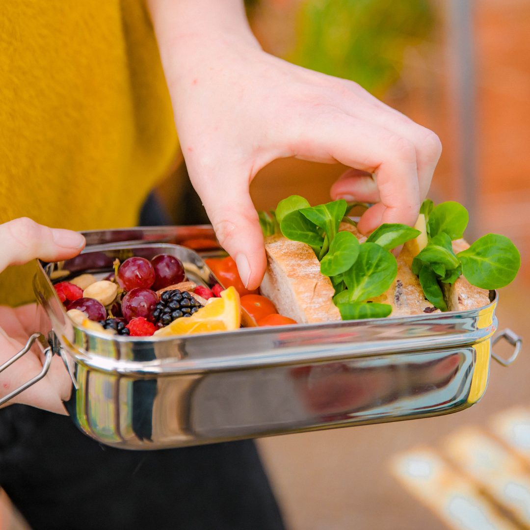 Jumbo 2 in 1 stainless steel lunch box from elephant box. box is open showing the inner snack pod containing fresh fruit and nuts. The main compartment has some chunky sandwiches & cherry tomatoes. someone is taking a sandwich out of the lunchbox.