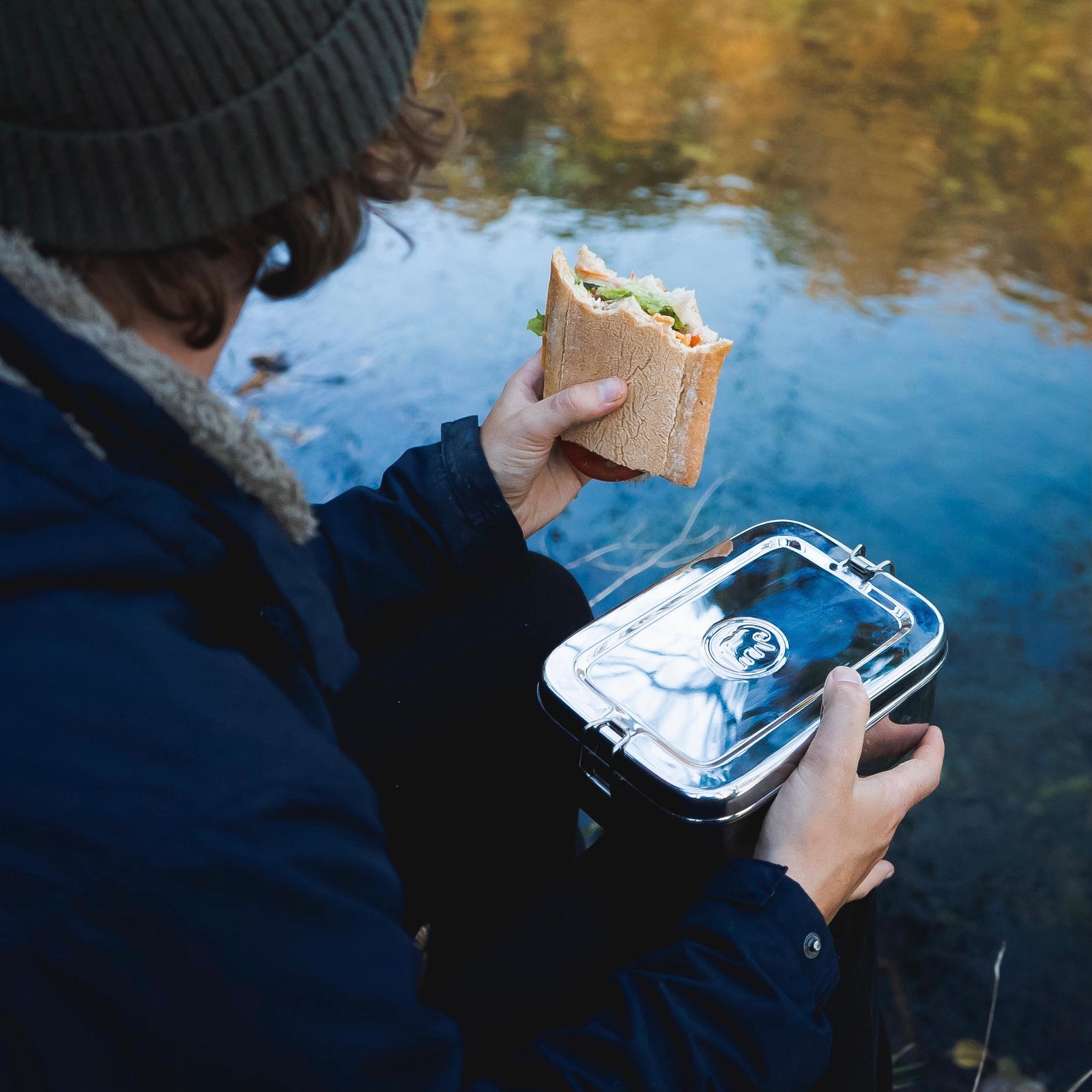 Person holding a sandwich and a lunch box by a body of water