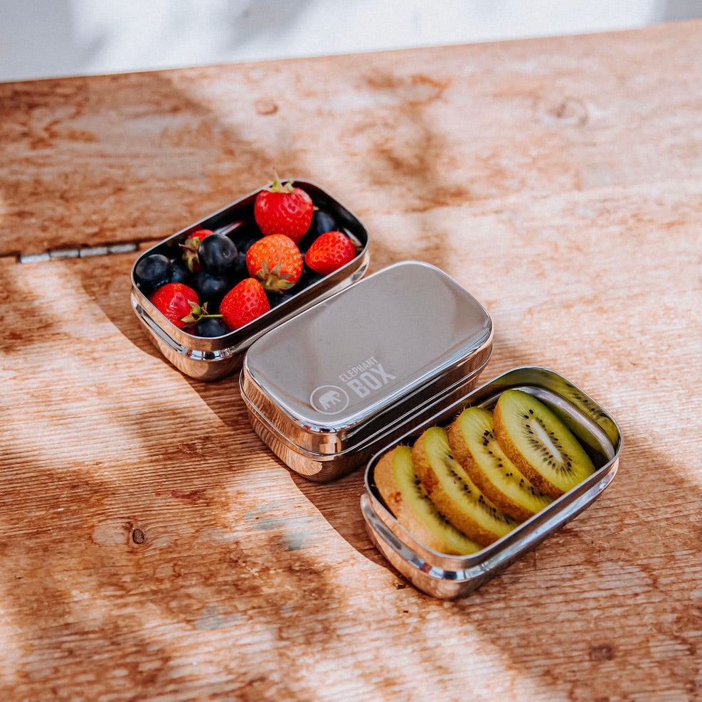 Three silver stainless steel snack storage containers with lids on a wooden surface one snack container has the brand name 'Elephant Box' on the top. and two are shown with mixed fruits inside.