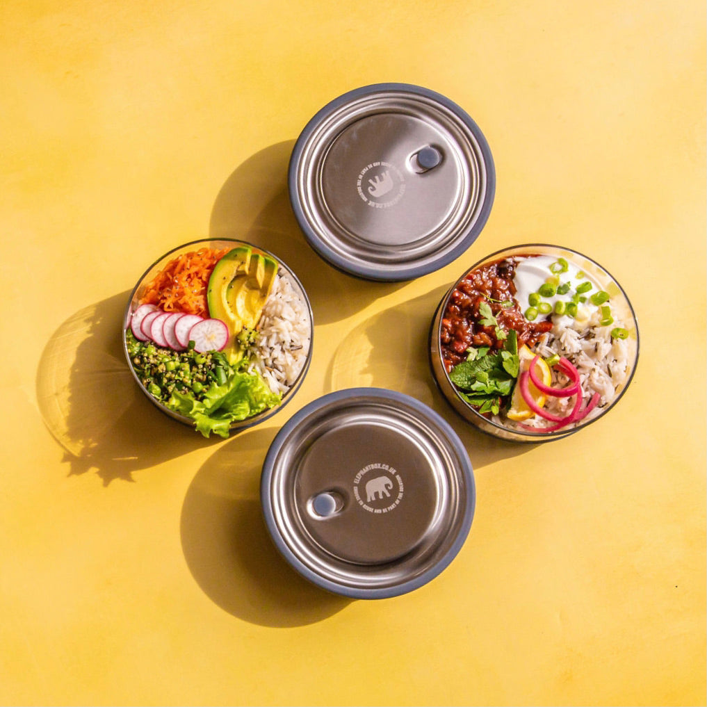 4 elephant box lunch bowls on yellow backgorund displaying a selection of meals. two of The glass lunch bowls have their stainless steel lids on. 