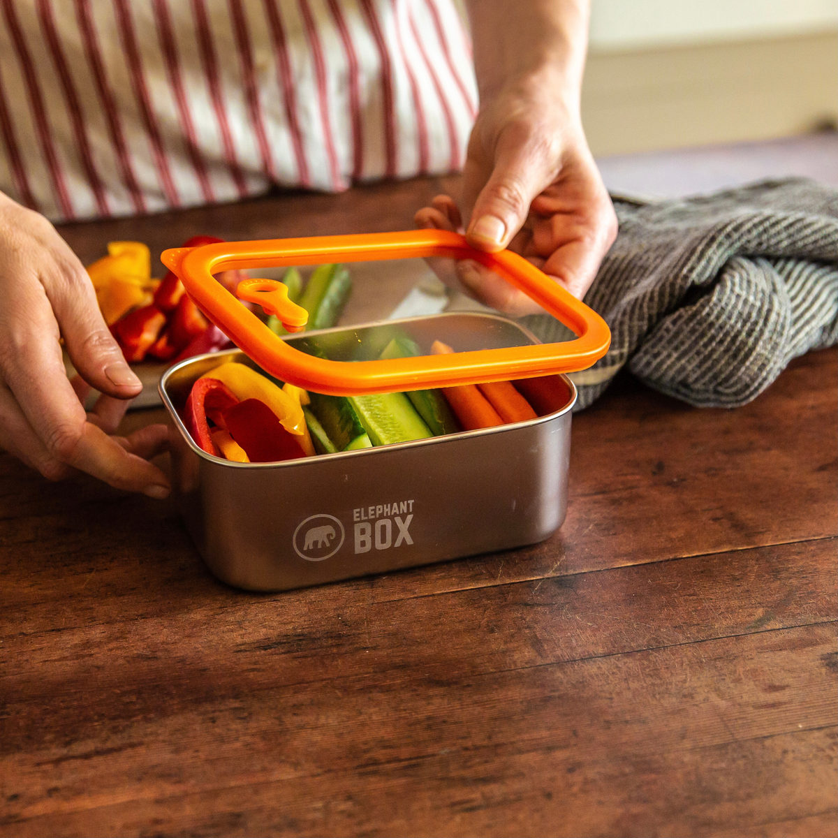 Person holding a stainless steel and glass  lunch box with colourful vegetables, branded 'Elephant Box', on a wooden surface.