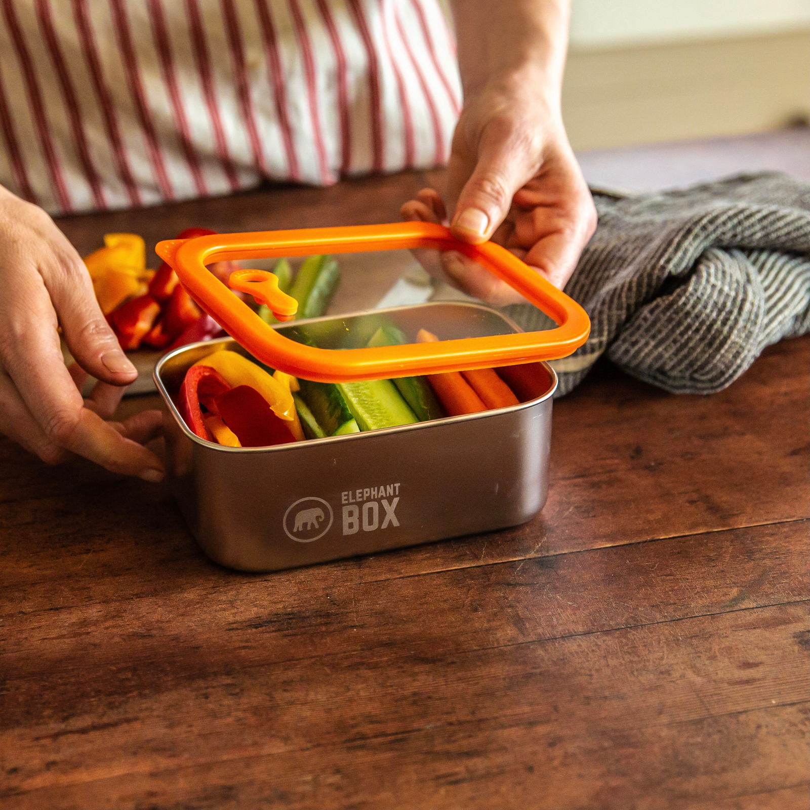 Person holding a stainless steel and glass  lunch box with colourful vegetables, branded 'Elephant Box', on a wooden surface.