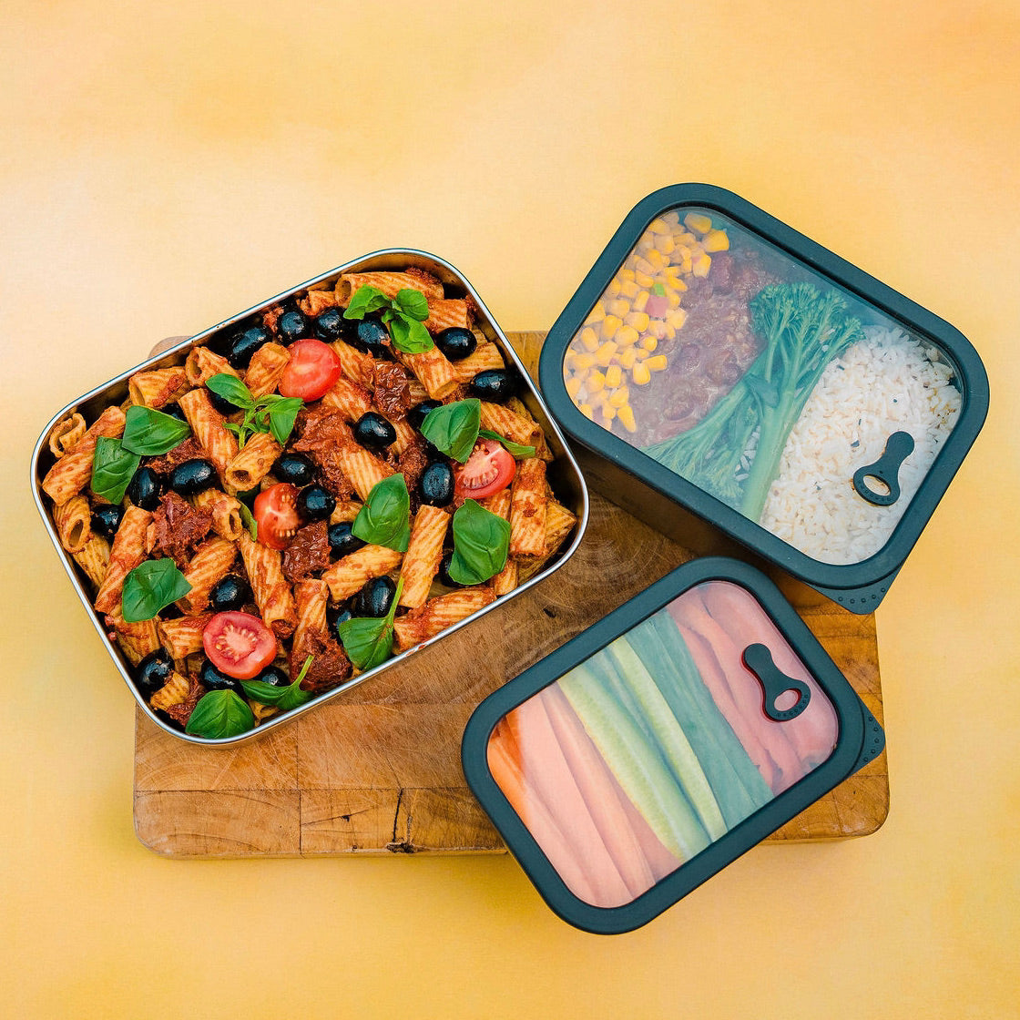 Three metal meal prep containers with different food items on a wooden board against a yellow background. The glass see through lid allows you to see the food. 