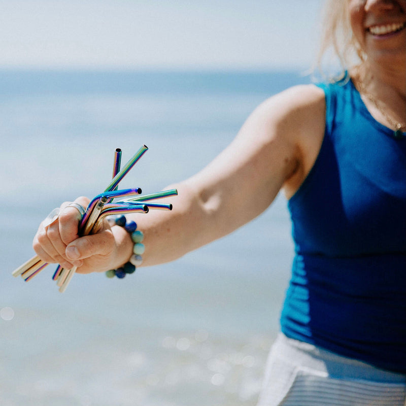 a person smiling and holding 4 bent multicoloured stainless steel straws in front of the ocean.