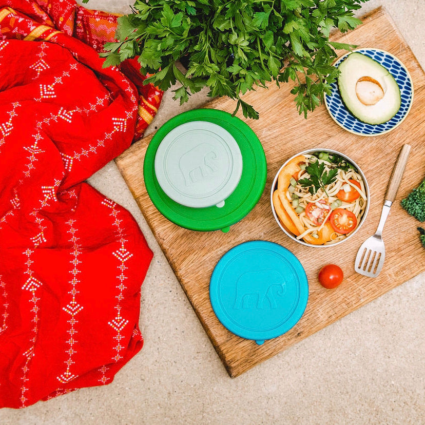 three round stainless steel food containers viewed from above on a wooden board showing food in one and the other two with their coloured silicone lids on top 