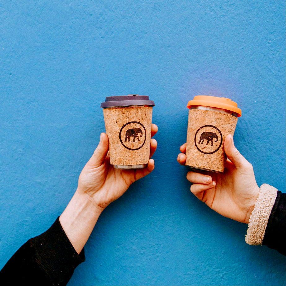 Two hands holding reusable to-go cups with a visible elephant logo on them, against a blue background.